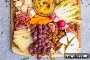 Overhead view of a colorful fruit and cheese platter featuring sliced brie, gouda, parmesan cubes, red grapes, hummus, olives, and crackers, beautifully arranged on a wooden board.