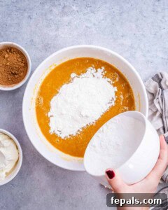 Spiced Pumpkin Cream Cheese Swirl Muffins 9 A hand gently pouring all-purpose flour over the wet pumpkin batter in a mixing bowl.