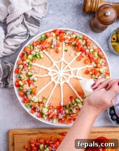 Top view of a hand piping a sour cream spider web design on top of the taco dip with diced veggies along the edges.