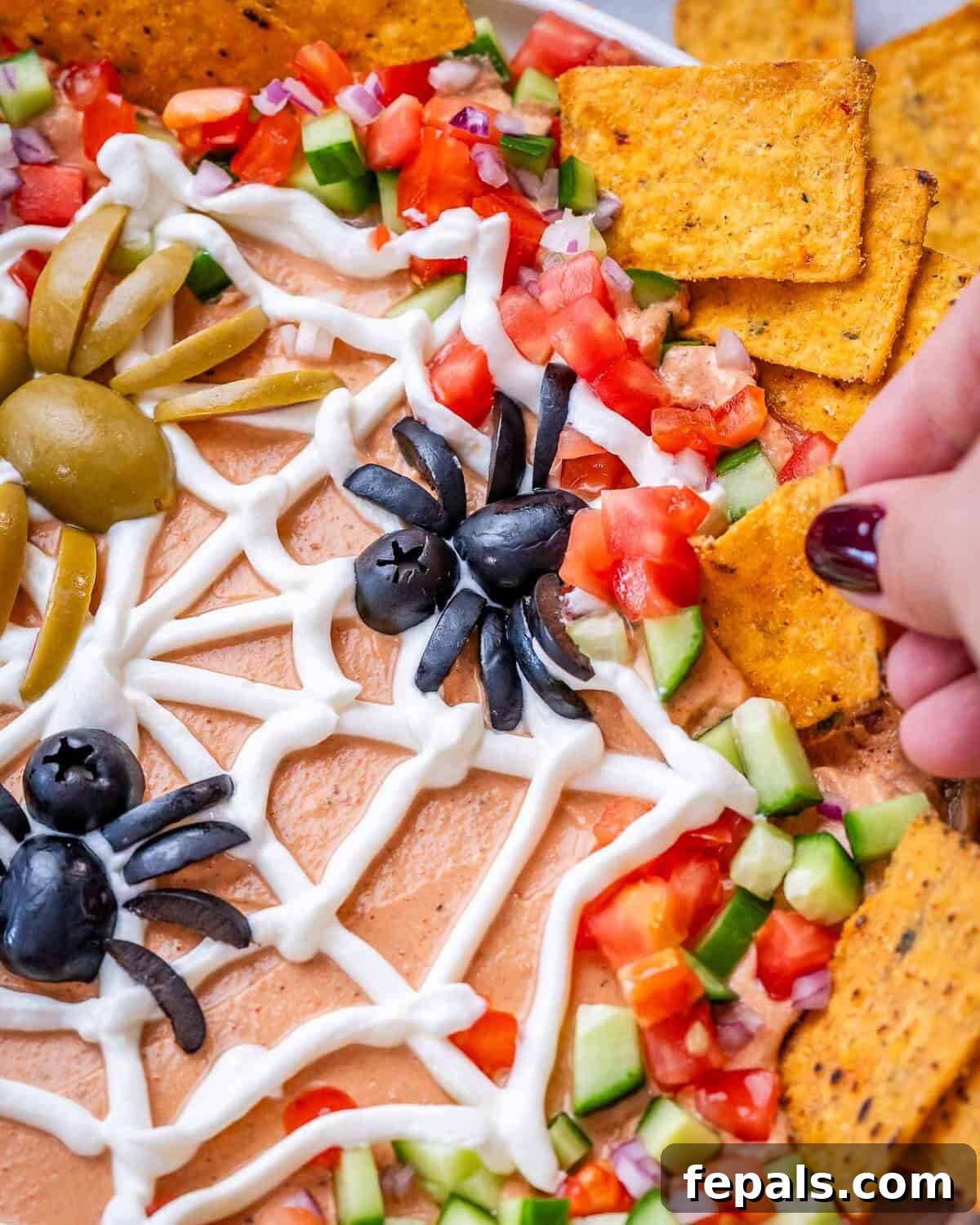 Hand grabbing a tortilla chip to scoop the dip out near a spider made out of olives on top of the sour cream spider web.