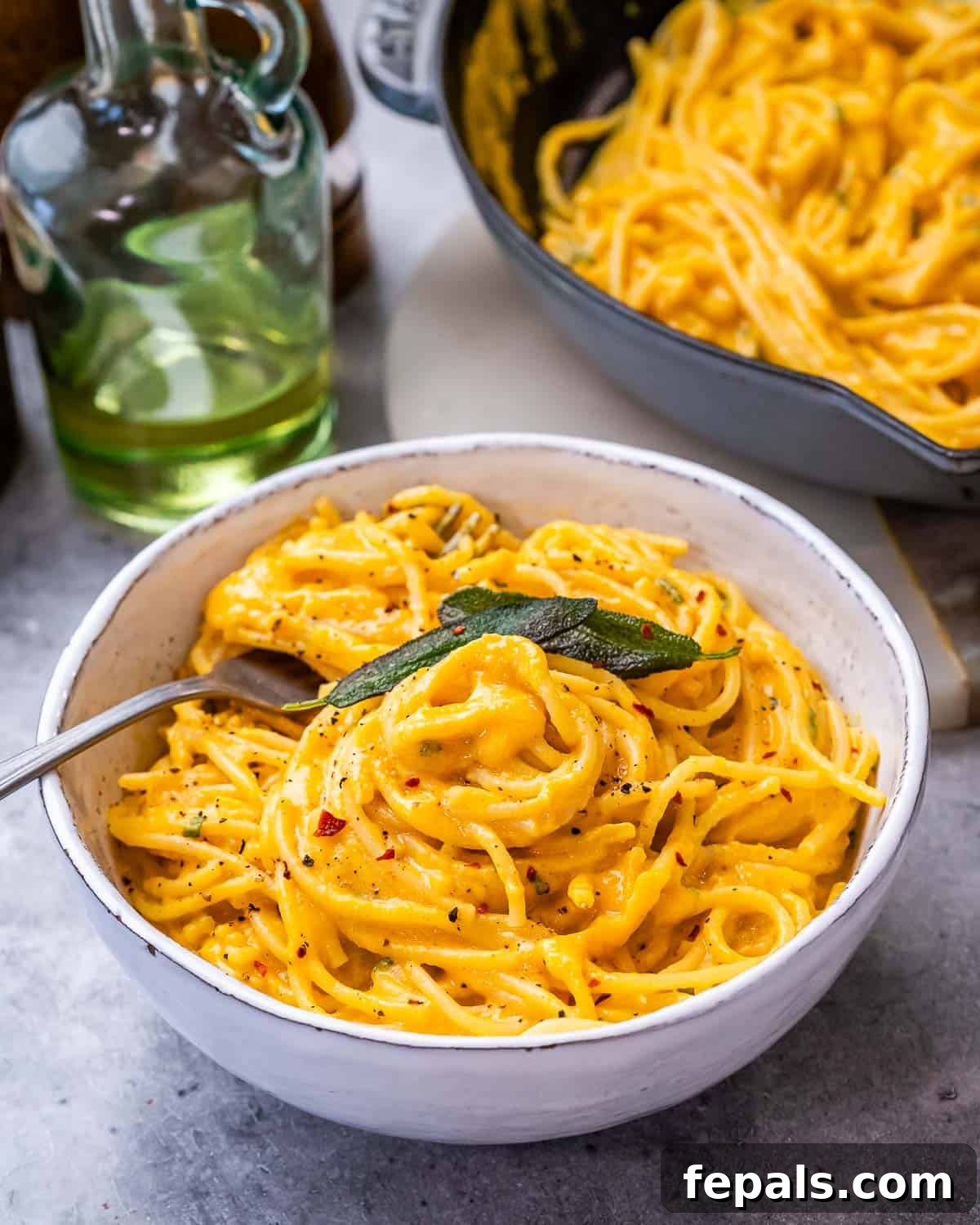 Simple Comfort Pumpkin Alfredo 13 A white ceramic bowl filled with creamy pumpkin alfredo pasta, with a cooking pan and a pumpkin in the soft-focus background.