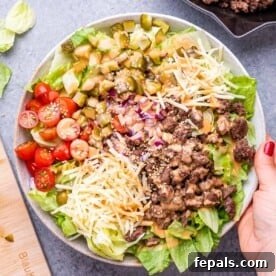 A hand holding a vibrant bowl of Big Mac Salad, playfully deconstructed with seasoned minced meat, fresh crisp lettuce, juicy tomatoes, and tangy pickles, all ready to be tossed.