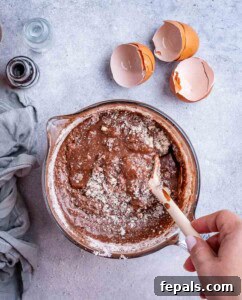 Mixing bowl with brownie batter being stirred using a wooden spoon, surrounded by cracked egg shells.