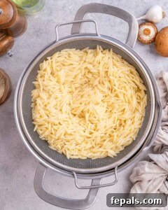 Cooked orzo draining in a metal strainer over a pot, ready to be added to the pan.