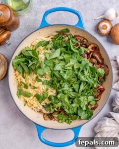 Overhead shot showing fresh baby spinach added to the pan of cooked orzo and mushrooms before mixing.
