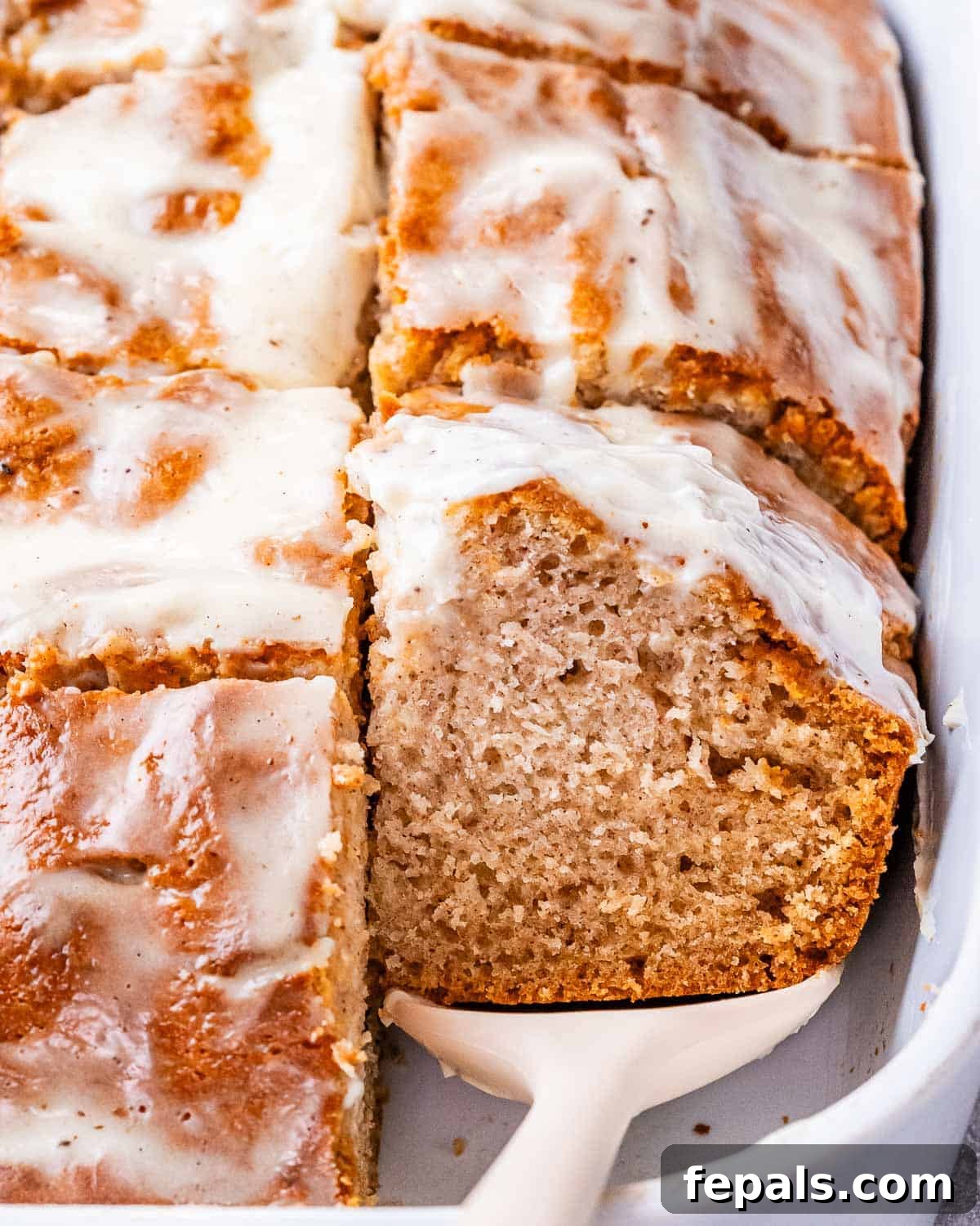 Cinnamon Swirl Dream Cake 7 A glorious close-up of a sliced cinnamon roll cake, richly coated with cream cheese glaze, resting in a white ceramic baking dish, showcasing its irresistible texture and design.