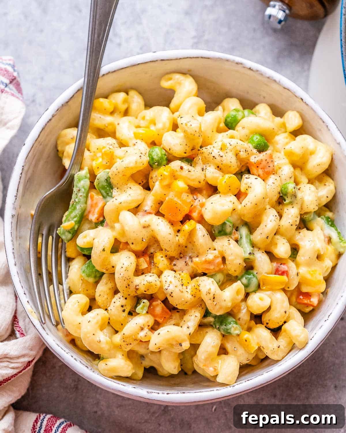 Vibrant Veggie Mac 2 A large white ceramic bowl filled with creamy veggie mac and cheese, featuring visible pieces of carrots, peas, and green beans. A fork is resting on the side of the bowl.