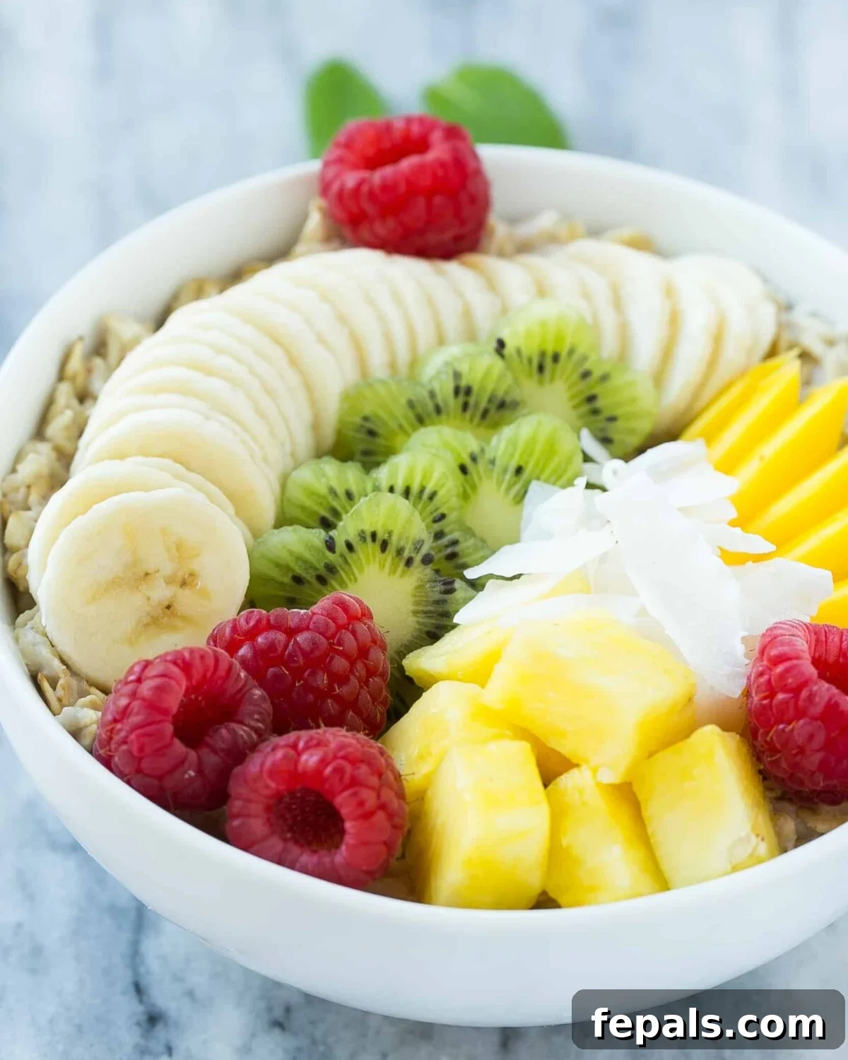 Vibrant Fruit and Oats Breakfast Bowl 2 A white bowl of creamy oatmeal generously topped with a vibrant assortment of fresh, sliced fruits including banana, kiwi, mango, pineapple, and raspberries, finished with a sprinkle of coconut flakes.