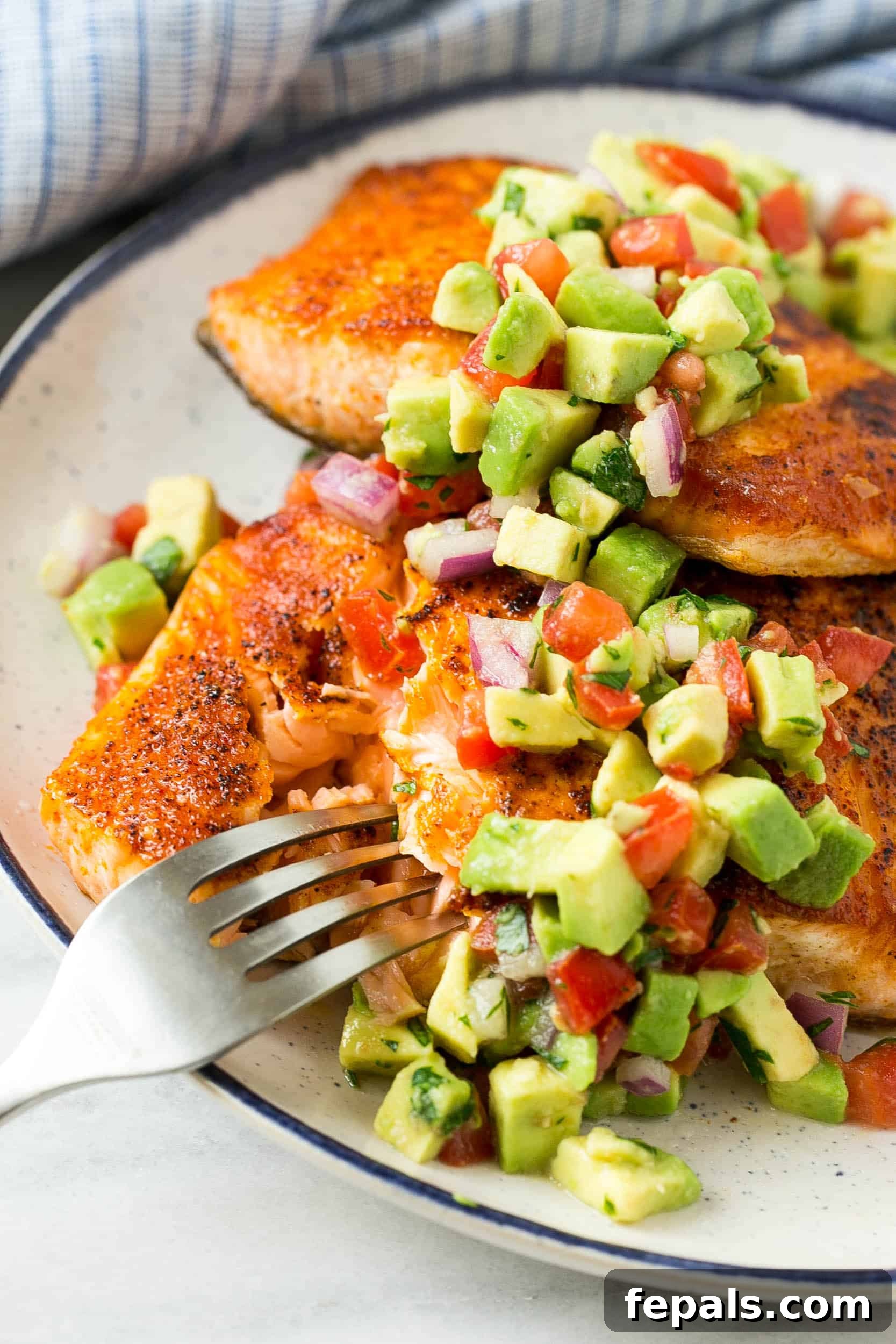 Perfectly fried salmon on a white plate, garnished with fresh avocado salsa and a fork ready to enjoy.