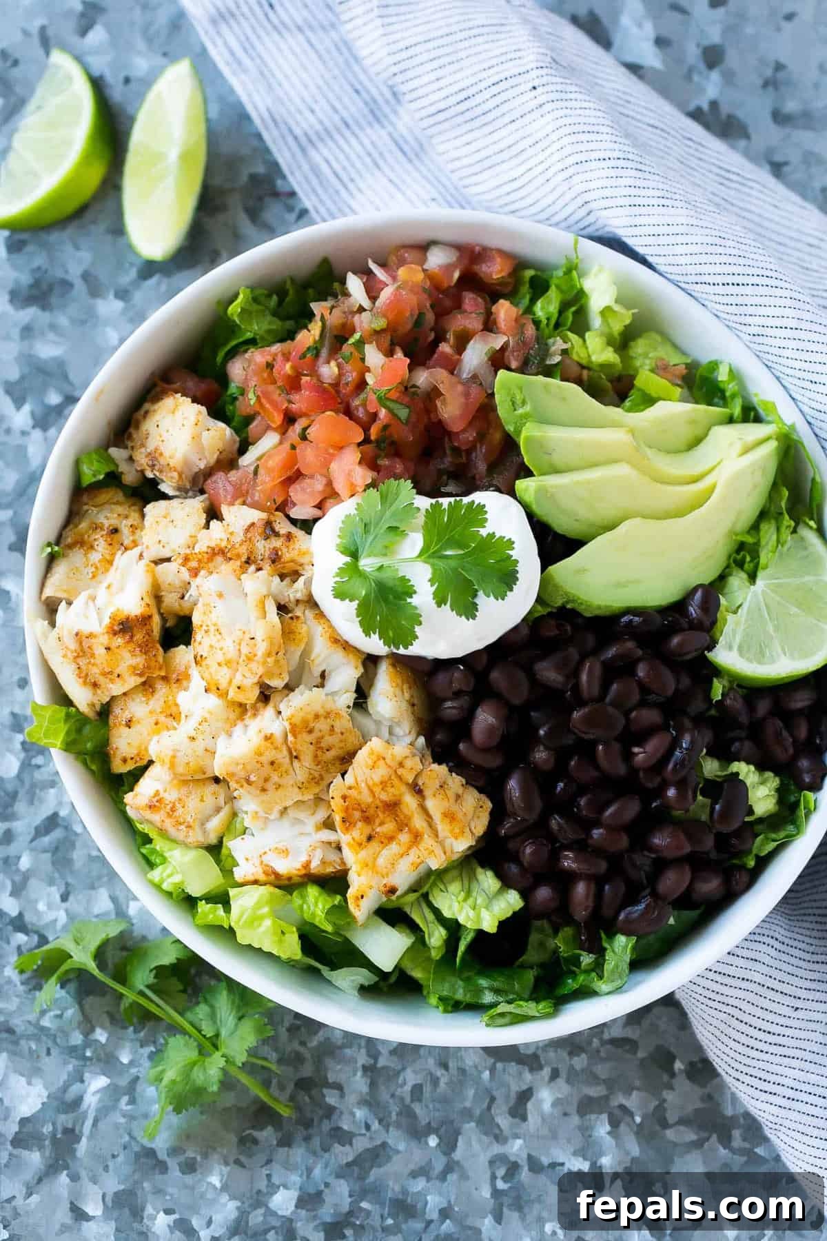 Fish Taco Salad Bowl Top view of a vibrant fish taco salad bowl, featuring crispy fish, black beans, avocado slices, sour cream, fresh cilantro, lime wedges, and salsa, all harmoniously arranged.