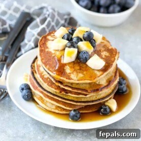 Blueberry Pancakes stacked on a round white plate with syrup and topped with chopped bananas and blueberries.