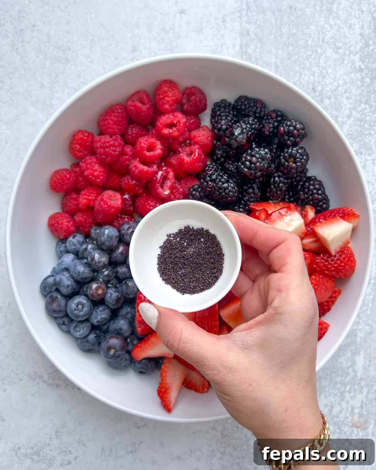 hand holding a small bowl of poppy seeds, sprinkling them over the berries.