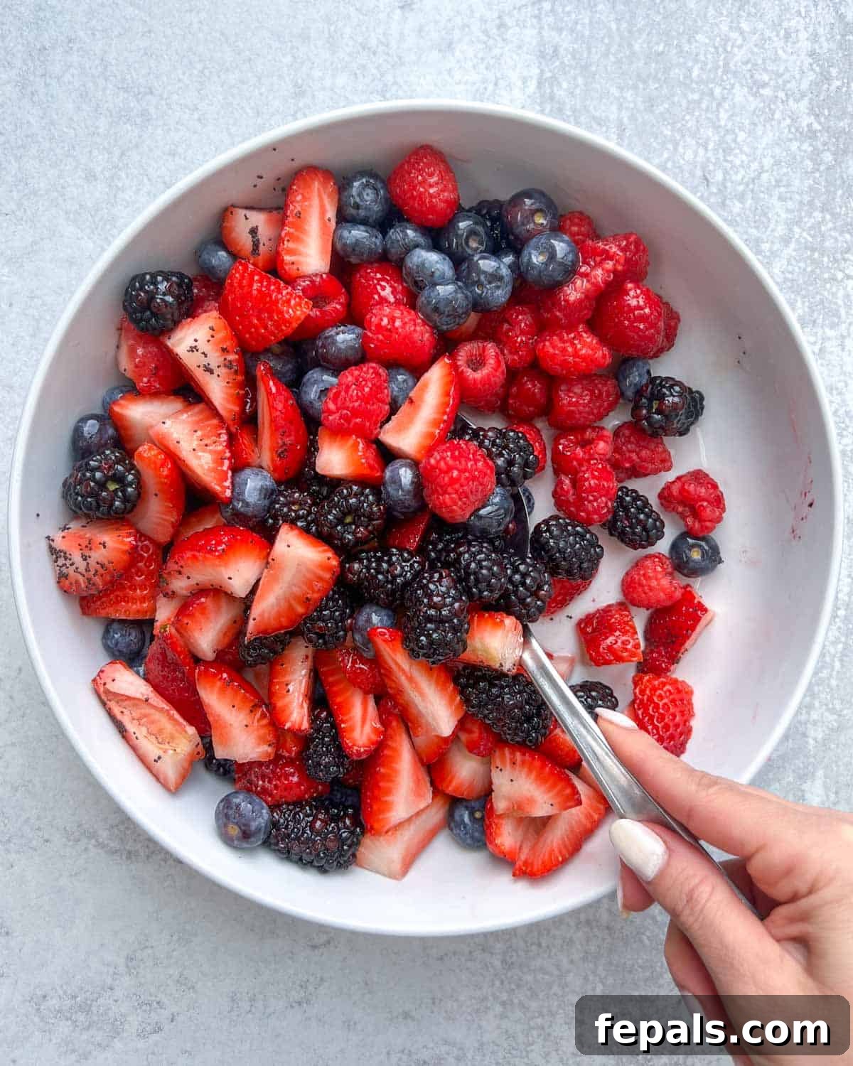hand mixing the berry salad using a spoon, ensuring an even coating of dressing.