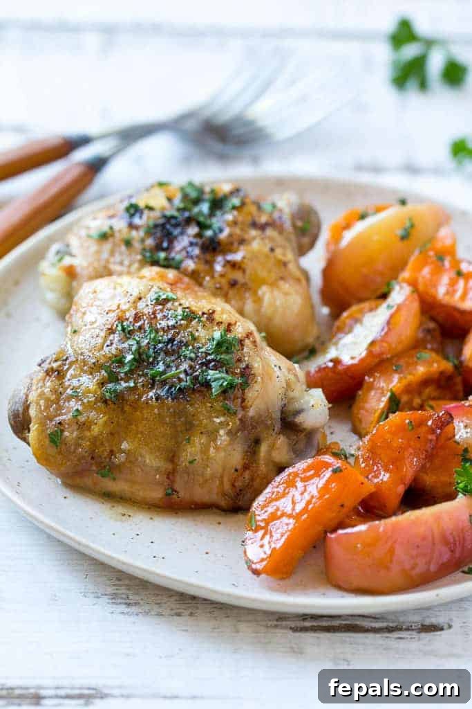 Close-up side view of a serving of roasted chicken thigh, maple sweet potatoes, and apples on a white plate