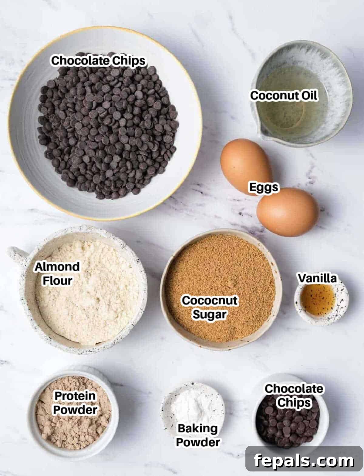 A selection of fresh ingredients including almond flour, chocolate chips, eggs, coconut oil, coconut sugar, and vanilla extract, laid out for making protein brownies.