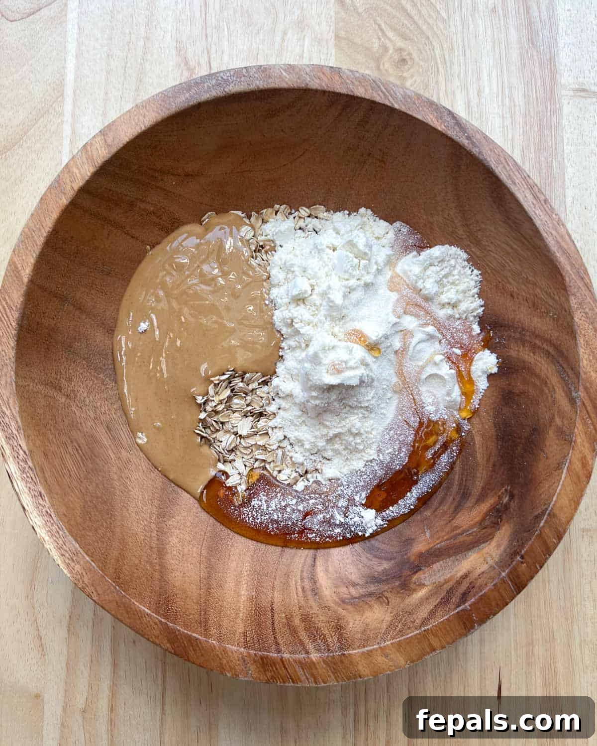 Oats, protein powder, peanut butter, and honey are added to a large wooden mixing bowl, ready to be combined.