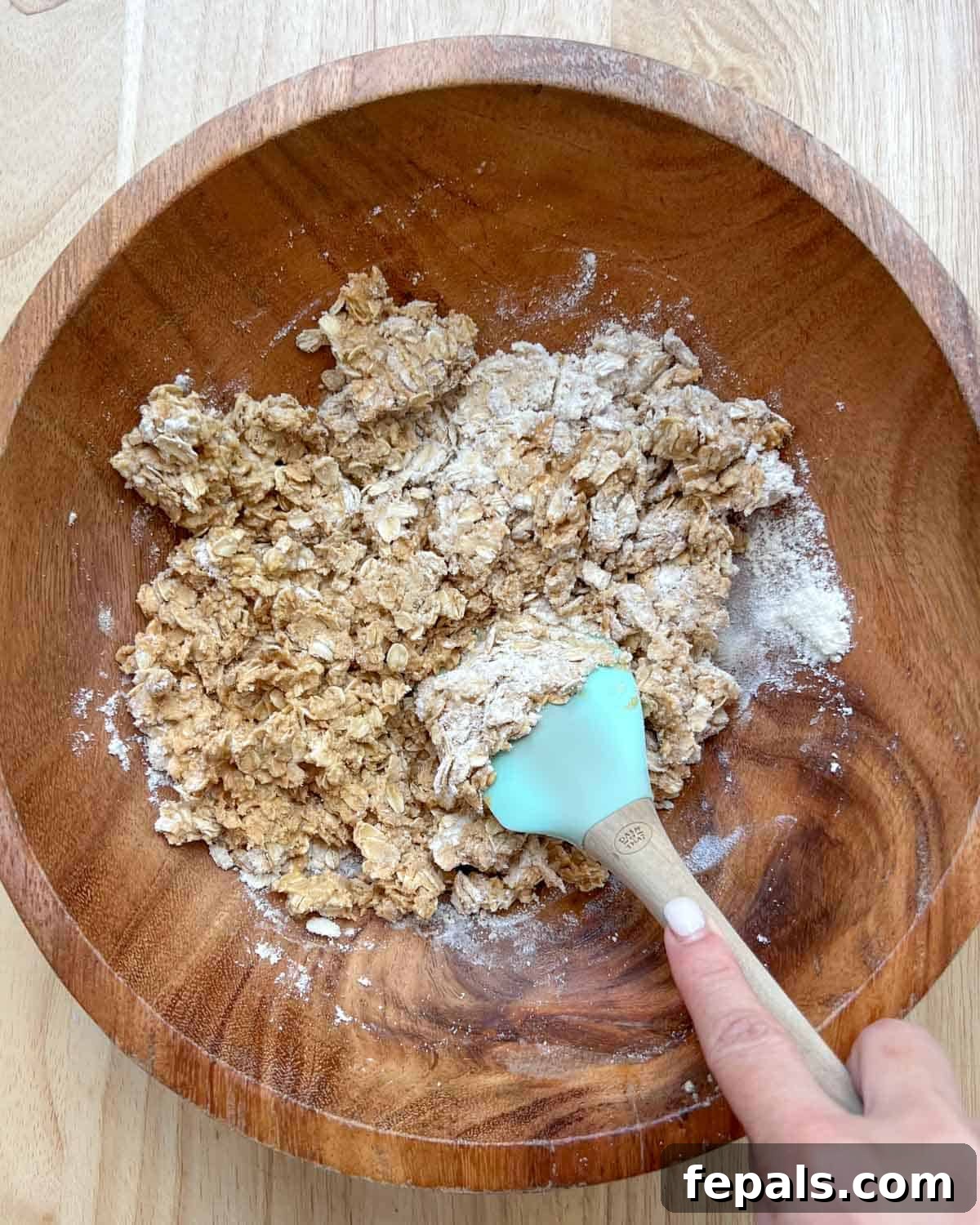A spatula is used to thoroughly mix the oats, peanut butter, honey, and protein powder in the bowl until a cohesive dough starts to form.