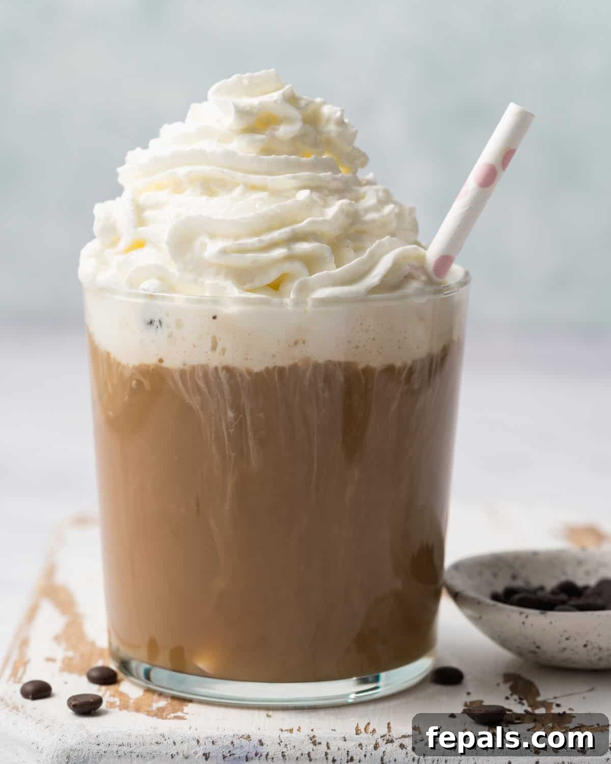 A generous dollop of whipped cream is being delicately added over the rich mocha protein shake in a glass cup, enhancing its dessert-like appeal.