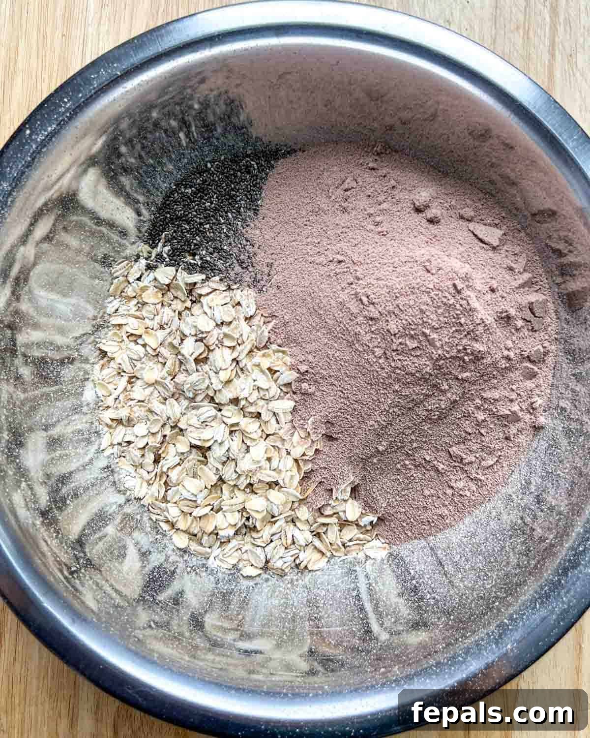 A stainless steel bowl filled with dry ingredients: rolled oats, chocolate protein powder, and chia seeds.
