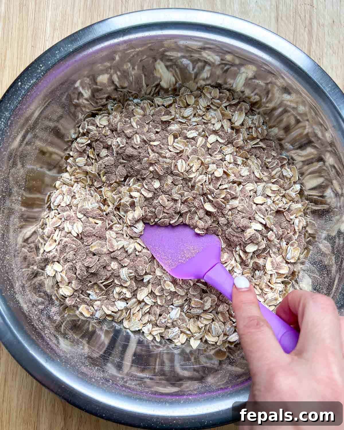 A hand using a purple spatula to thoroughly mix the dry ingredients together in a bowl.
