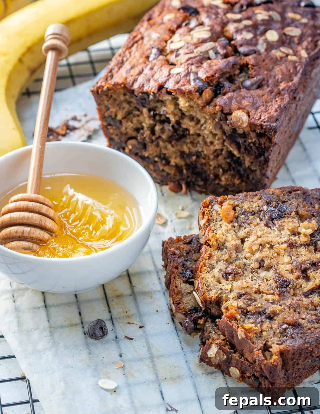A whole oatmeal chocolate chip banana bread loaf on a cooling rack, next to a bowl of honey, with some slices already cut.
