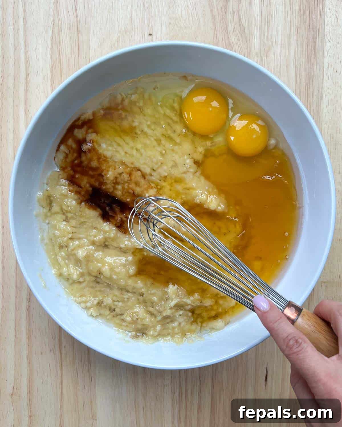 Eggs, mashed bananas, vanilla extract, honey, and coconut oil mixed in a white bowl with a whisk.