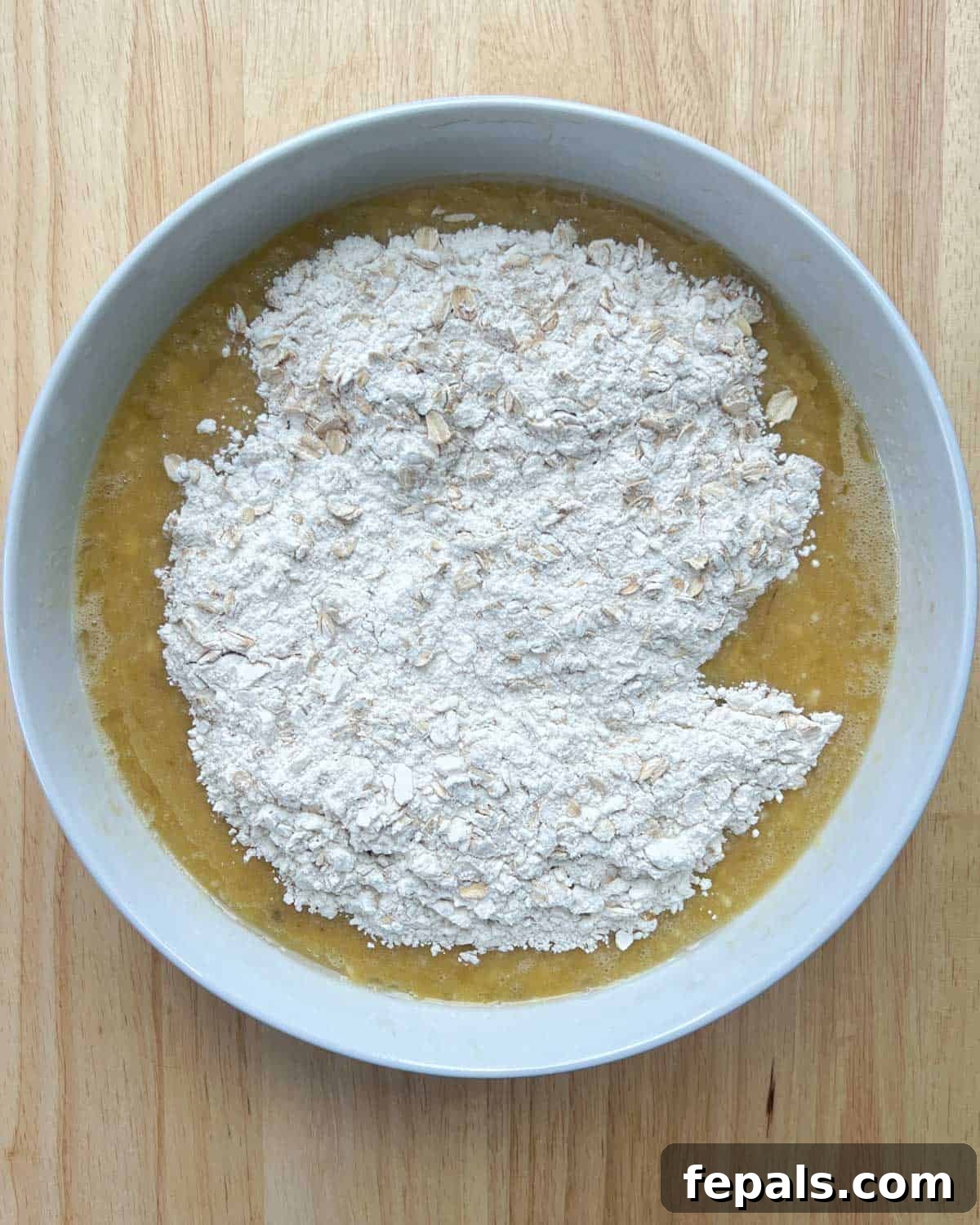 Dry flour ingredients being added over the wet egg and banana mixture in a round bowl, ready for whisking.