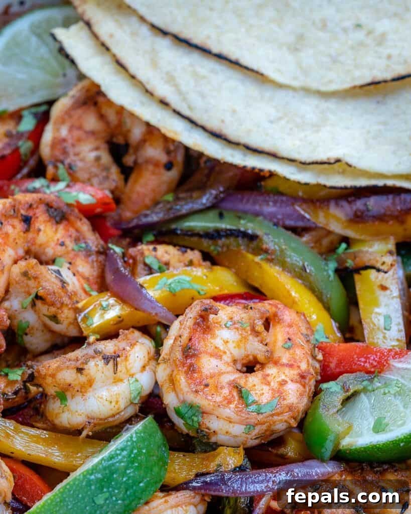Close-up image of a cast-iron pan full of cooked shrimp fajitas, with warm flour tortillas stacked beside it, ready for serving.