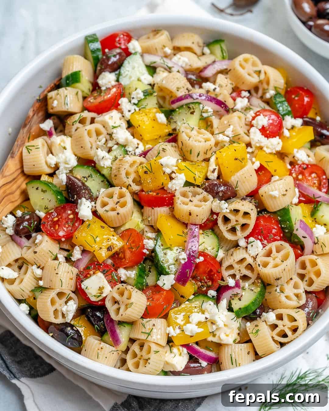 Zesty Greek Pasta Salad 2 A beautifully arranged Greek pasta salad in a round white bowl, brimming with colorful vegetables and crumbled feta cheese.