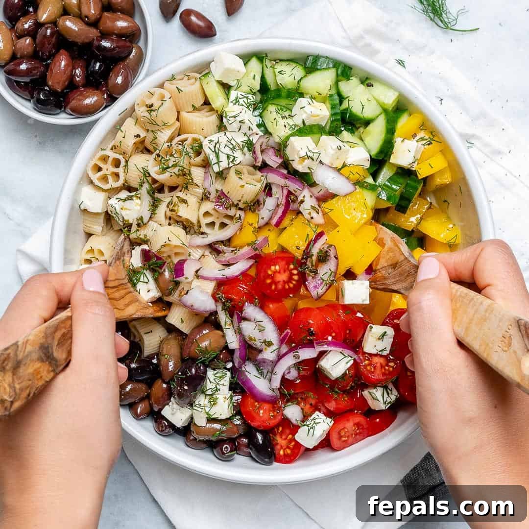 Zesty Greek Pasta Salad 3 A hand gently mixing a colorful Greek pasta salad in a large round bowl with a wooden spoon and fork, showcasing its fresh ingredients.