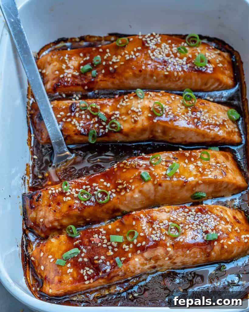 Sticky Teriyaki Salmon Bake 3 Metal spatula lifting a piece of perfectly baked teriyaki salmon from a white dish, garnished with green onions and sesame seeds