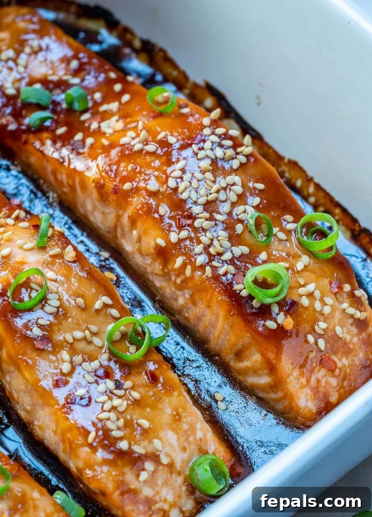Sticky Teriyaki Salmon Bake 4 Close-up side view of baked teriyaki salmon in a white dish, with sauce at the bottom and garnished with sliced green onions and sesame seeds
