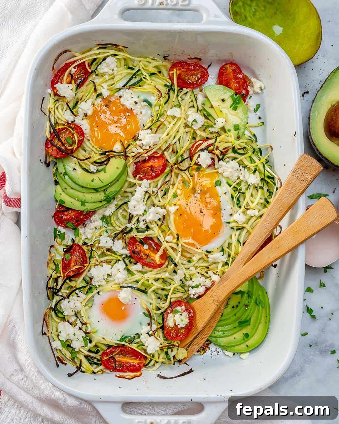 Zoodles in a baking dish with baked eggs and cherry tomatoes, topped with sliced avocados.