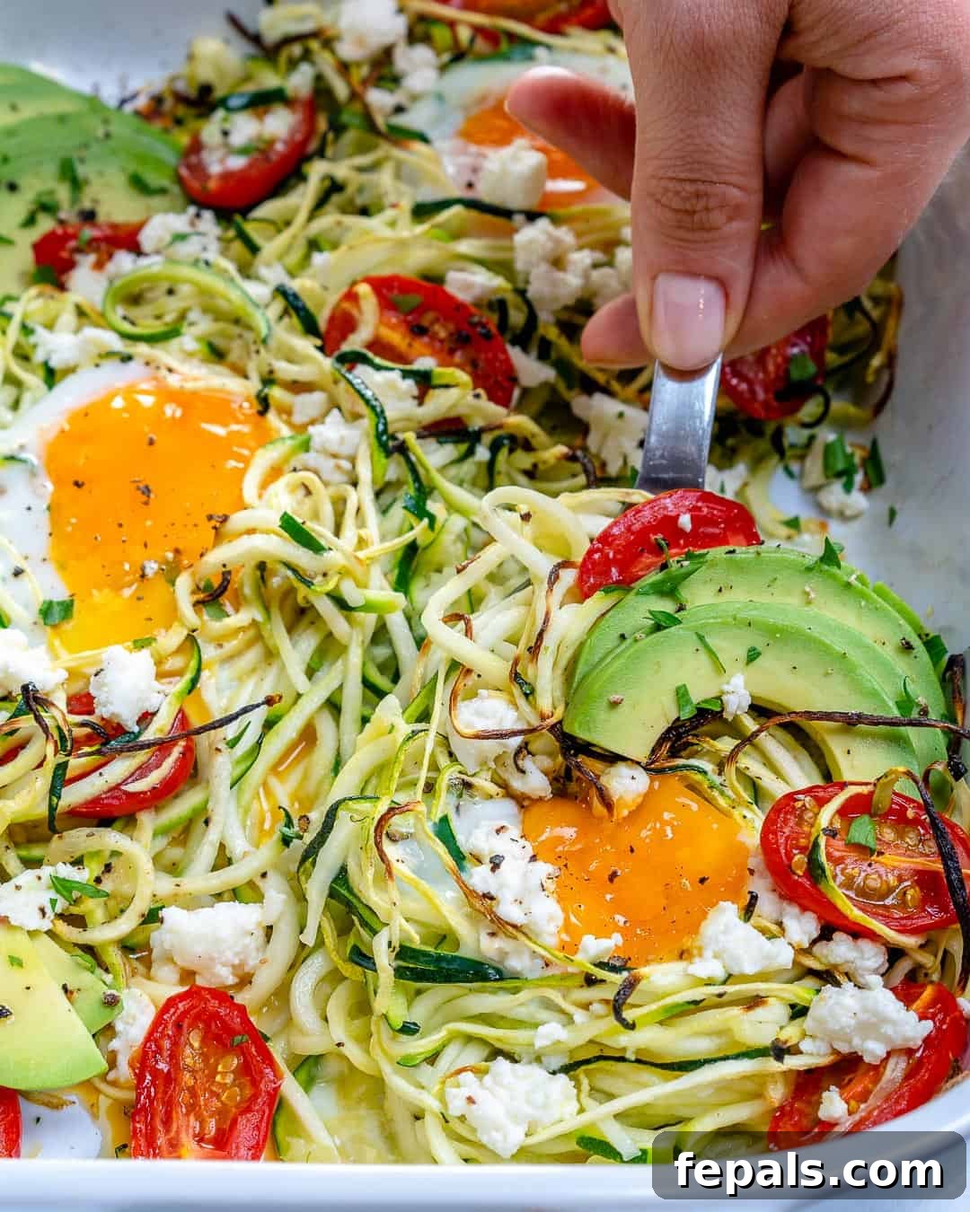 Hand grabbing a portion of baked eggs with zoodles in a dish.
