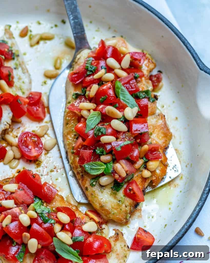 Spatula picking up a Pesto grilled chicken breast from a pan topped with pinenuts and chopped tomato mix.