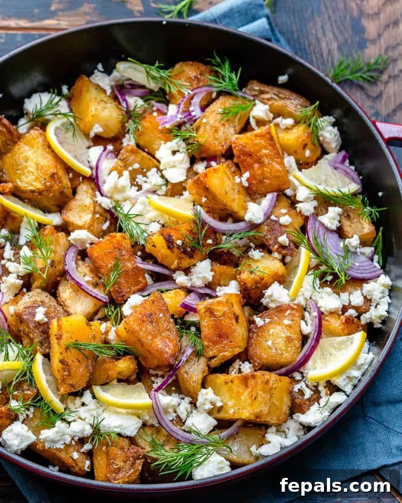 Zesty Greek Roasted Potatoes 2 top view of a black skillet with crispy potatoes topped with crumbled feta cheese, lemon garnishes, fresh dill, and sliced red onions