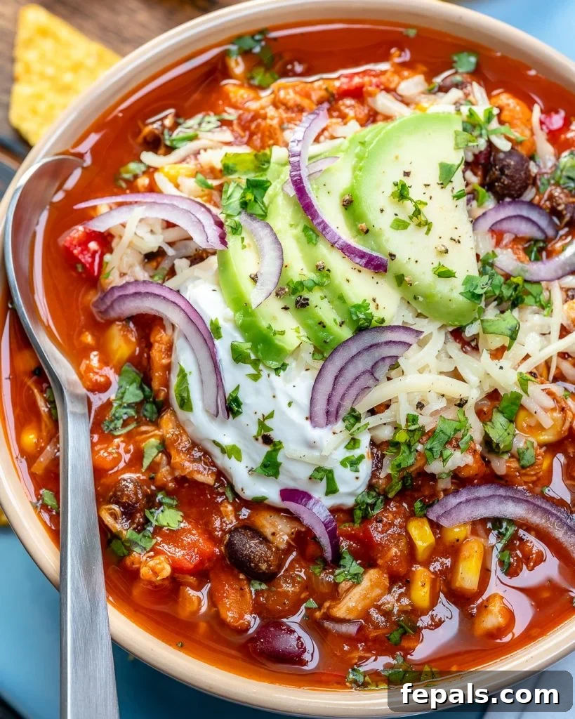 Ultimate Chicken Chili 2 Top view of a white bowl with red looking chicken chili with spoon in bowl, topped with sliced avocado, sour cream, and shredded cheese