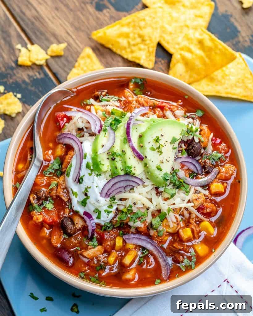 Ultimate Chicken Chili 8 a white round bowl that has red chicken chili topped with shredded cheese, sliced purple onions, avocado, and sour cream