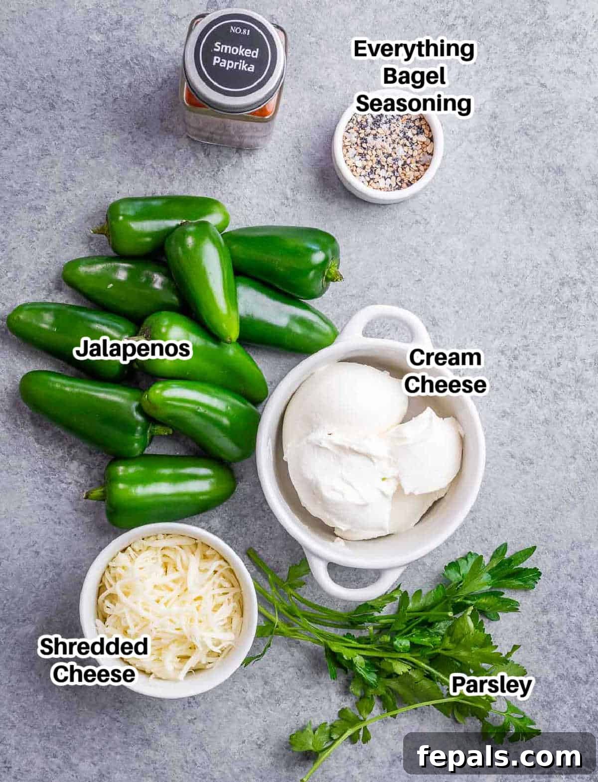 A selection of ingredients laid out on a wooden board, including fresh green jalapenos, blocks of cream cheese and cheddar, and small bowls of spices like everything bagel seasoning and smoked paprika, ready for making jalapeno poppers.