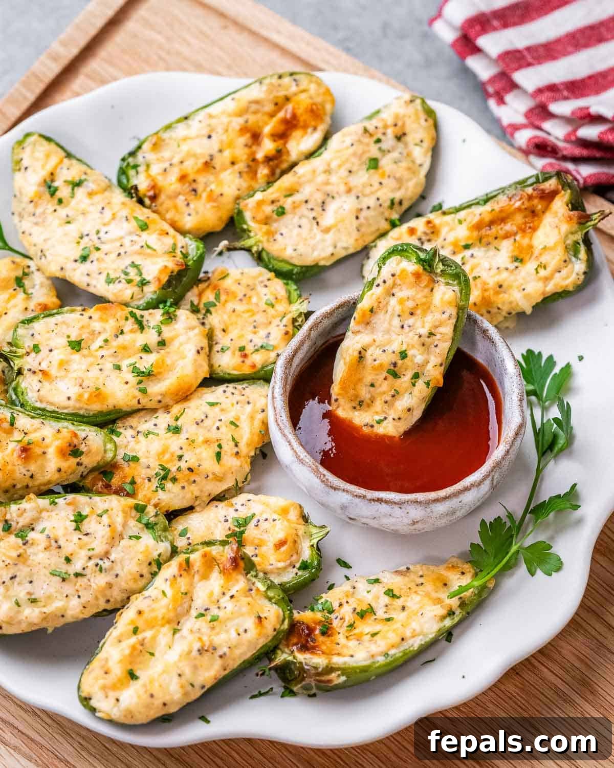A plate with beautifully baked jalapeno poppers, one of which is half-dipped into a small bowl of red sauce, highlighting its creamy filling.