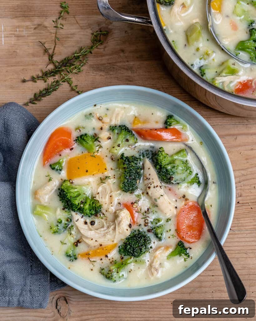 Velvety Chicken and Broccoli Potage 4 top view of a white round bowl of a creamy chicken and broccoli soup with carrots and yellow bell peppers, with spoon on the right side