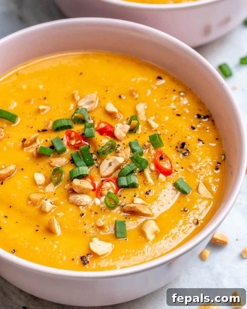 close up shot of an orange looking soup in a bowl topped with sliced green onions, chili peppers, and peanuts 