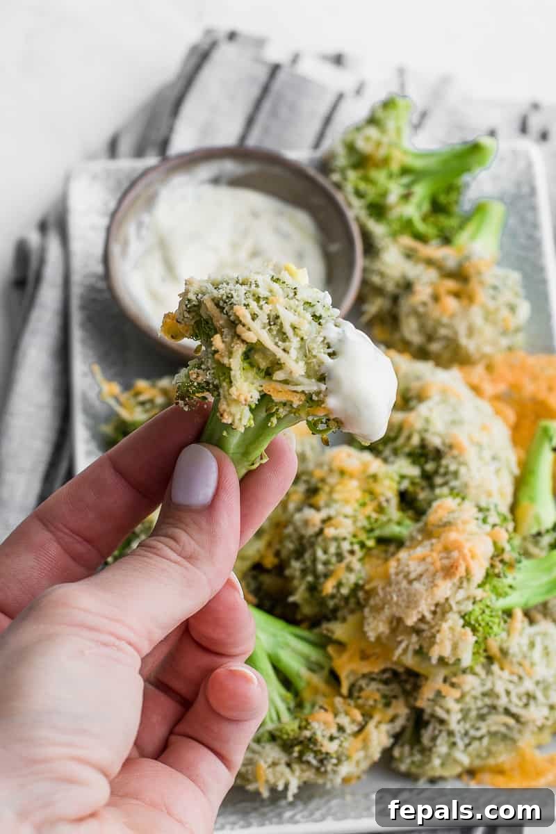 A hand holding a single cheesy baked broccoli floret over the serving plate, showcasing the crispy coating.