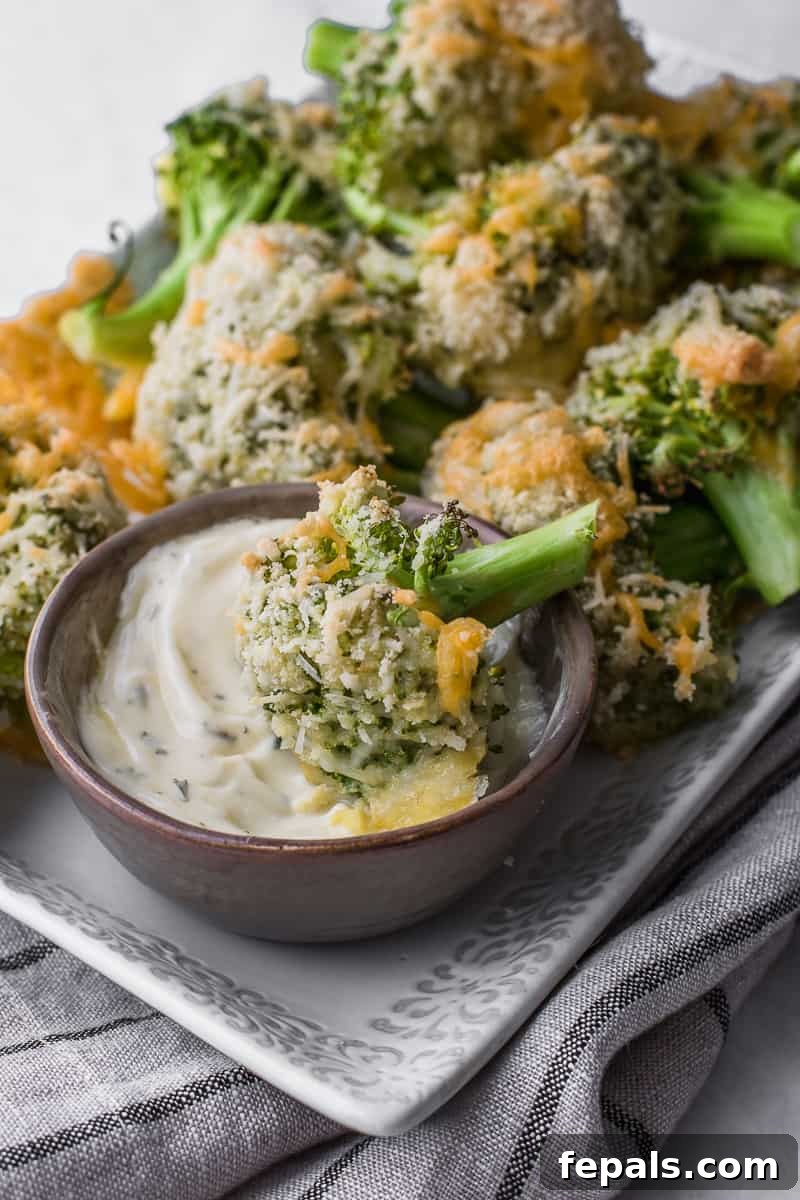 Side view of baked broccoli on a serving plate with one floret being dipped into a small bowl of white sauce.