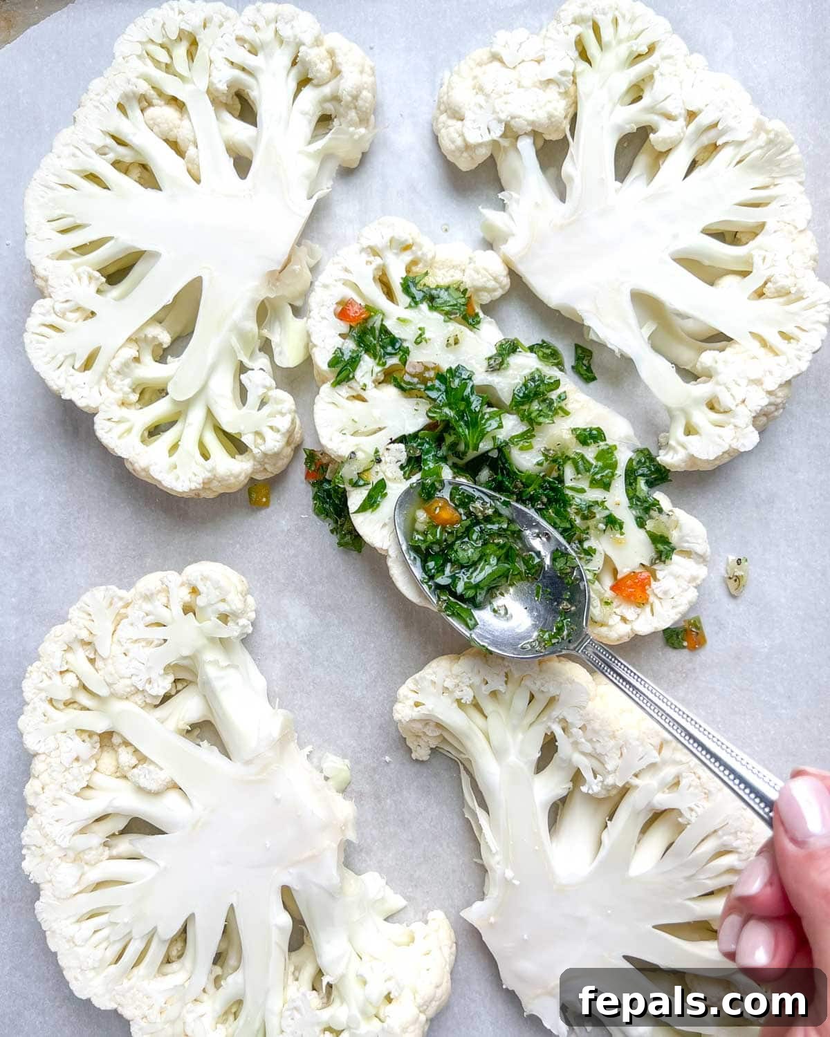 Chargrilled Cauliflower Steaks with Fresh Chimichurri 7 A hand using a spoon to drizzle generous amounts of chimichurri sauce over cauliflower steaks on a baking sheet.