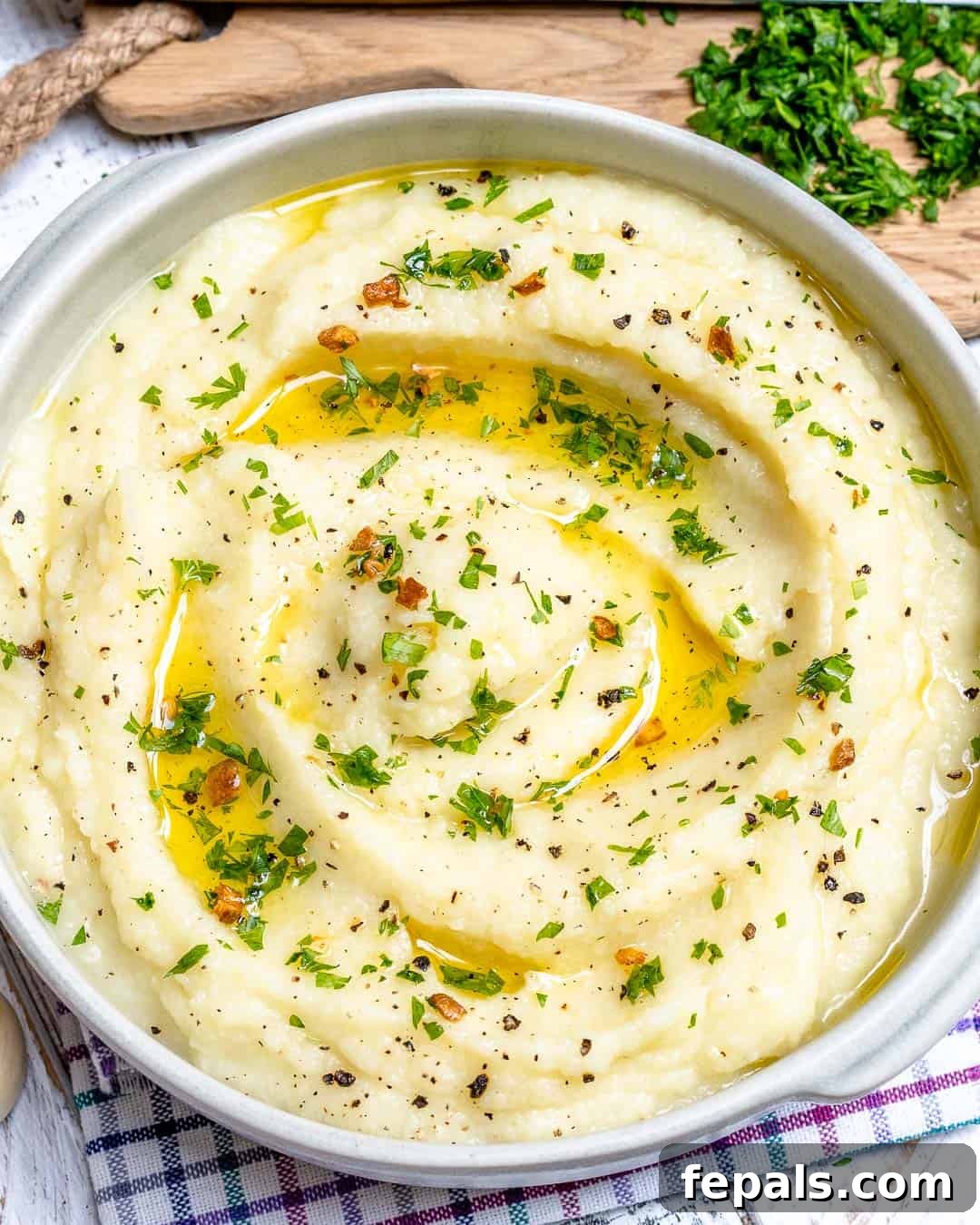 Whipped Garlic Butter Cauliflower 2 A round white bowl filled with creamy garlic butter mashed cauliflower, topped with a generous dollop of melted butter and sprinkled with fresh green herbs. The texture is smooth and inviting.