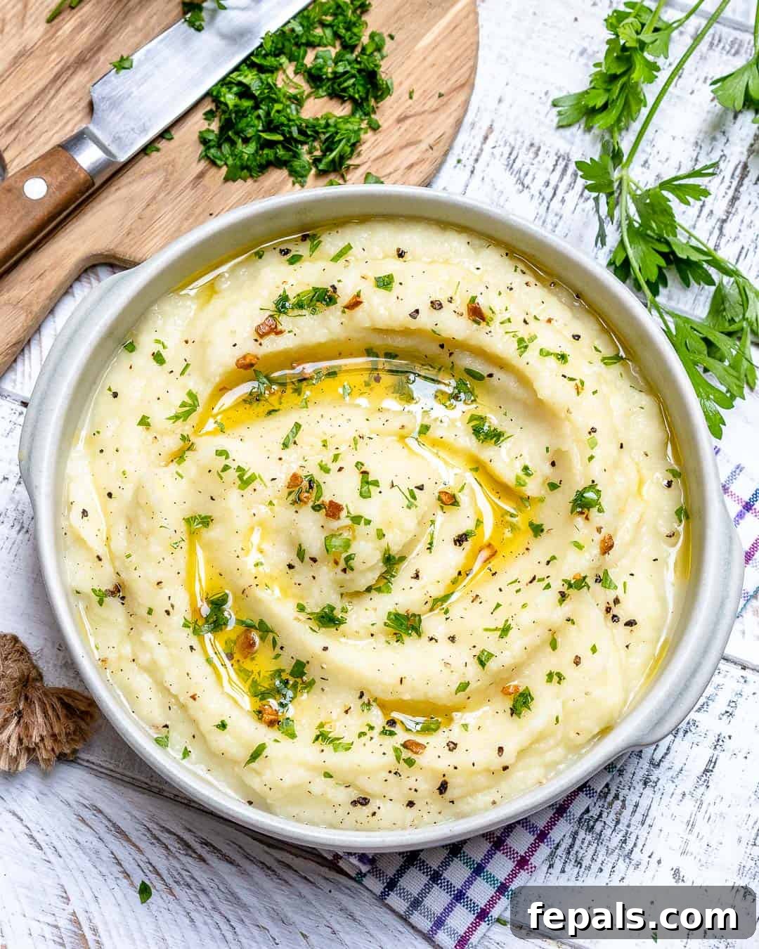 Whipped Garlic Butter Cauliflower 5 A bowl of creamy mashed cauliflower positioned next to a wooden cutting board, with fresh herbs nearby, highlighting the freshness of the ingredients.