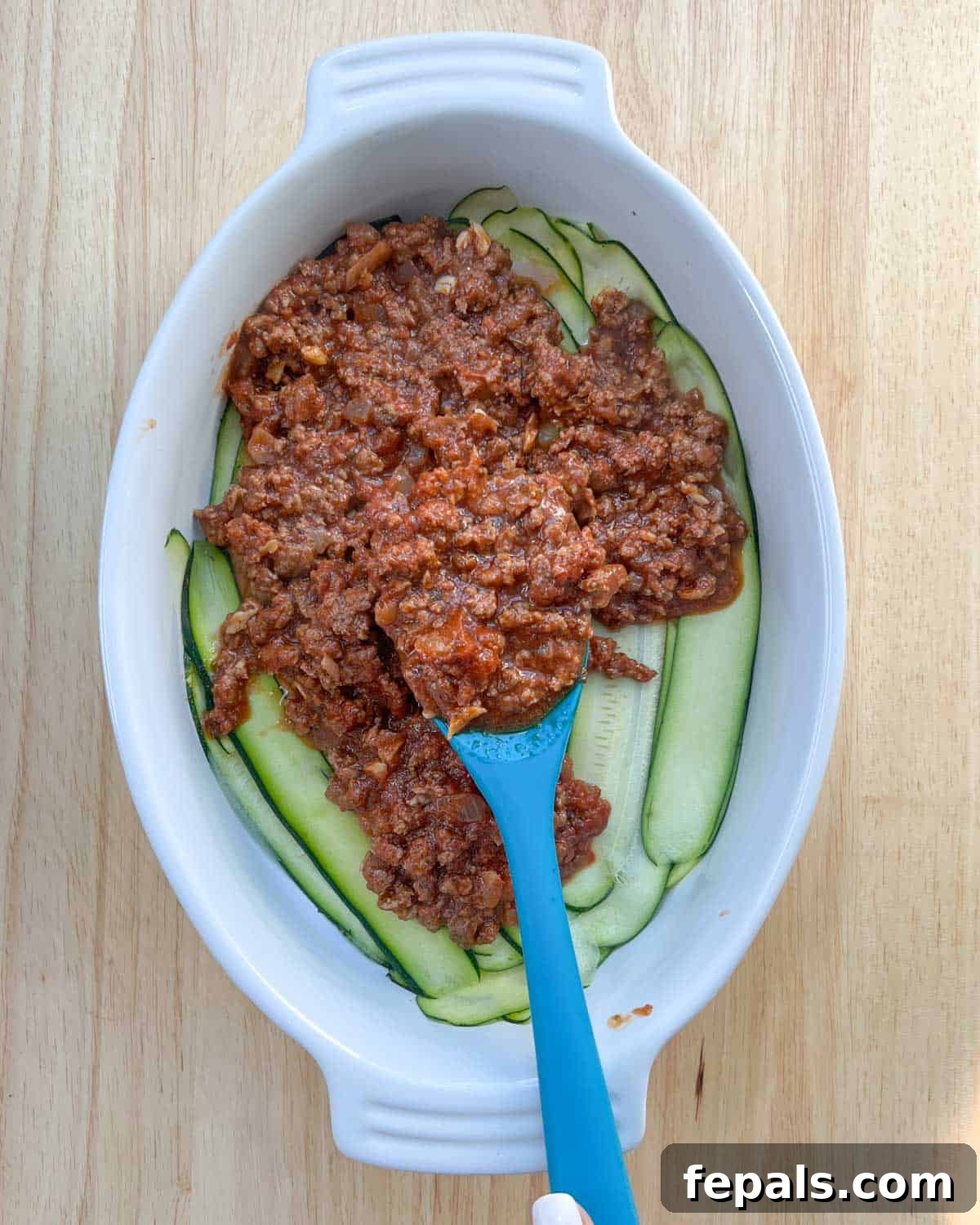Satisfying Zucchini Lasagna Bake 6 The initial layer of thinly sliced zucchini lining the bottom of a baking dish, with a spoonful of rich meat sauce being spread evenly over it, beginning the lasagna assembly.