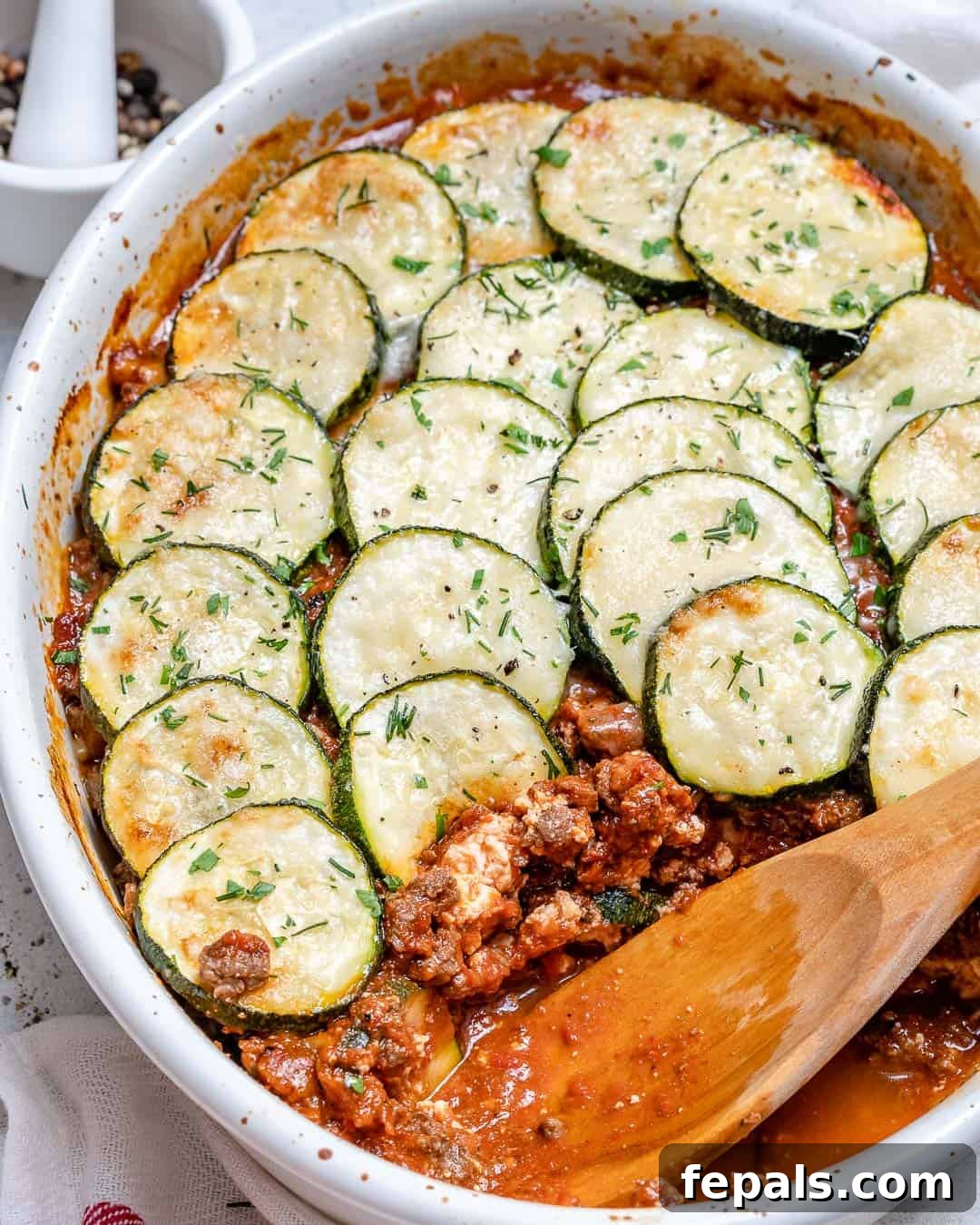 Savory Greek Zucchini Moussaka 5 A wooden spoon serves a portion of low-carb moussaka, showcasing layers of zucchini and meat sauce, garnished with fresh herbs.