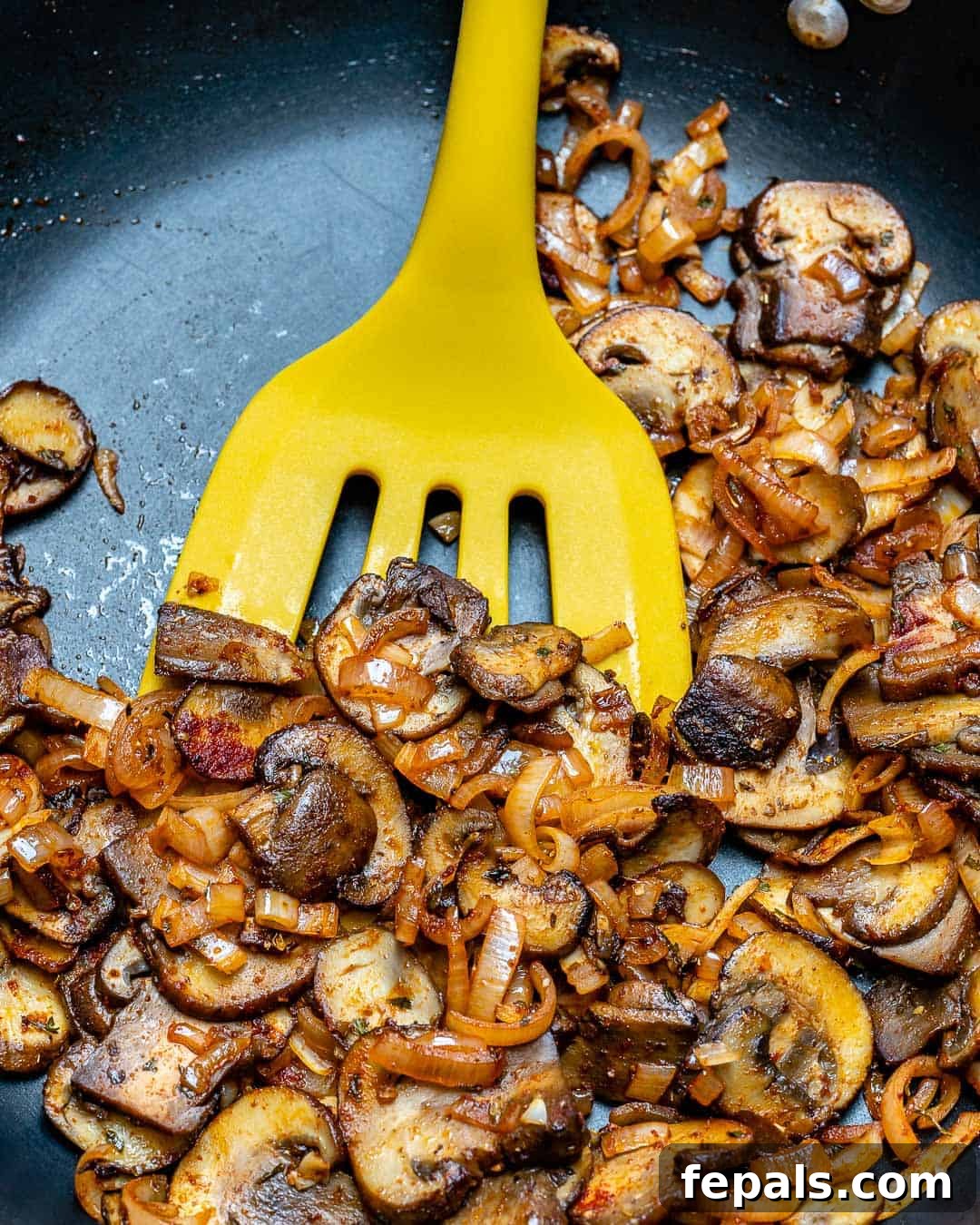 A yellow spatula stirring a vibrant mix of sautéed mushrooms and onions in a skillet, preparing the aromatic base for the casserole.